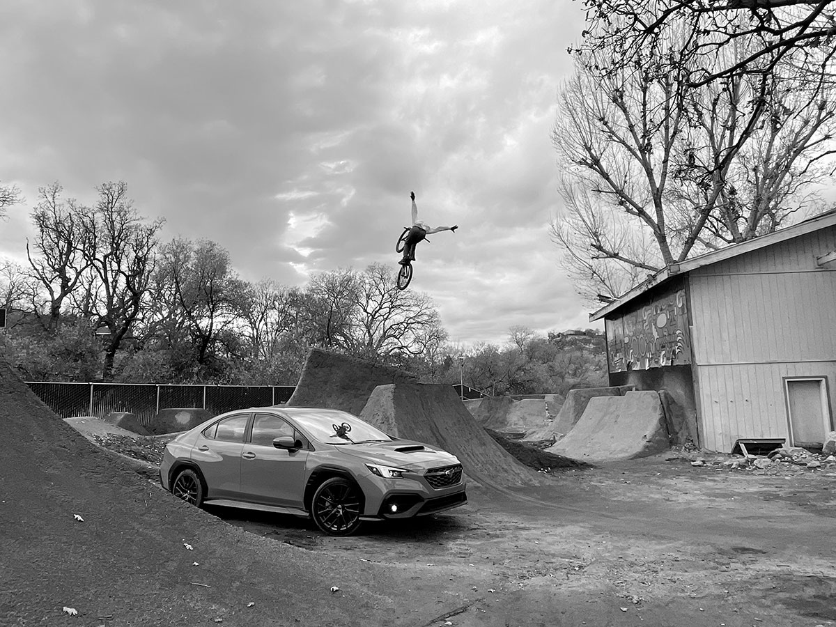 BMX rider catching air over dirt jumps next to a Subaru WRX